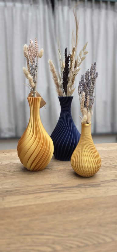 Drie vazen in verschillende kleuren met gedroogde bloemen, op een houten tafel.